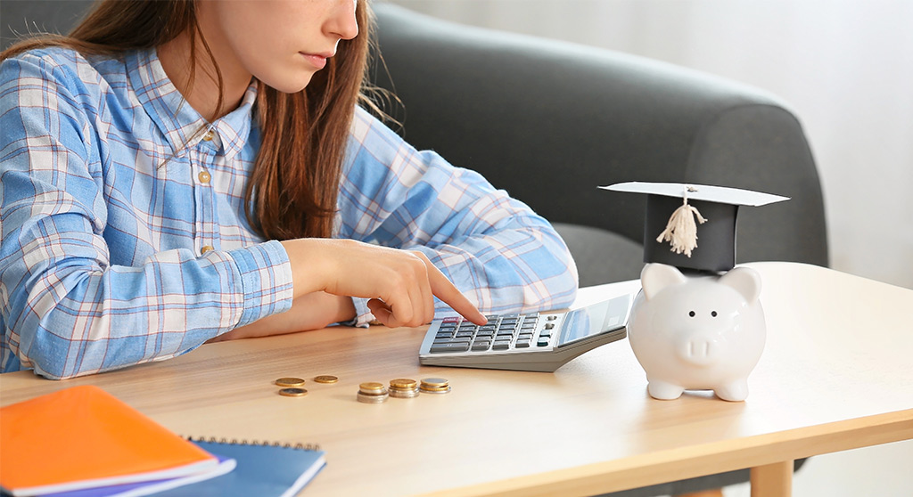 young adult looking at calculator with a piggy bank with a graduation cap